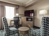 Hotel room with two patterned chairs, a round table, a desk, an office chair, a lamp, and a TV on a cabinet. Light from a window in the background.