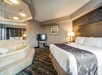 Hotel room with a king-sized bed, jacuzzi, wall-mounted TV, and a lamp on a bedside table. Neutral color palette with patterned carpet and mirrored wall panels.