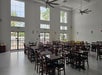A spacious dining hall with rows of empty wooden tables and chairs, high ceilings, large windows, ceiling fans, and natural light coming in.