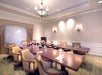 A large, empty conference room with a long wooden table, beige chairs, a chandelier, and decorative lighting. The walls are patterned and a window is partially covered by drapes.