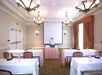 A small conference room with tables and chairs, a projector on a table, and a screen at the front. Chandelier lights hang from the ceiling.