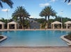 Outdoor swimming pool surrounded by palm trees and cabanas under a bright blue sky.