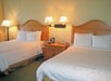 Two neatly made beds with wooden headboards, white linens, and pillows in a hotel room. A bedside table with a phone and lamp is between the beds. Wall lamps provide additional lighting.