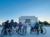 A group of people with bicycles pose in front of the Lincoln Memorial at dusk. Some wear masks and helmets.