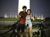 Two people wearing helmets stand with their arms around each other and smile at the camera, with bicycles in front of them and the Washington Monument in the background at night.