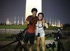 Two people with helmets and backpacks stand smiling near bicycles, with a tall illuminated monument in the background.