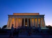 The Lincoln Memorial at dusk, illuminated with yellow and orange lights, with visitors gathered on the steps and silhouetted against the evening sky.
