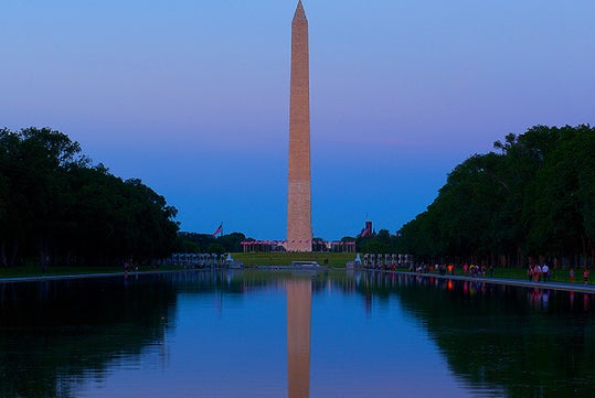 The Washington Monument reflected in the Lincoln Memorial Reflecting Pool at dusk, with trees lining both sides and a clear sky overhead.