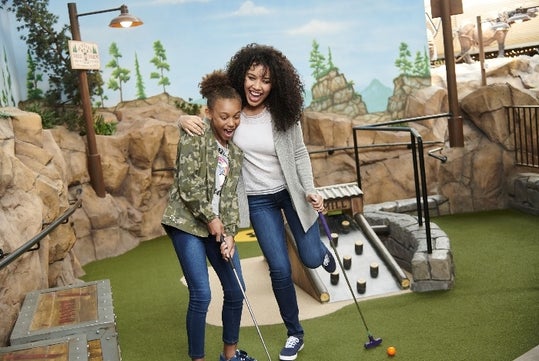 A young and middle age women smiling and laughing while playing mini golf with a wall of rock behind them at Moose Mountain Adventure Golf at Mall of America.