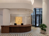 Modern hotel lobby with a curved wooden reception desk, two laptops, pendant lights, patterned tile floor, and a potted plant near glass entrance.