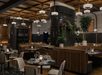 Modern restaurant interior with round tables, wooden chairs, dark tiled pillars, hanging lights, and plants. The atmosphere is cozy and elegant.