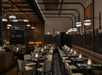A dimly lit, elegantly furnished restaurant with neatly arranged tables and chairs, modern overhead lighting, and dark wood walls.