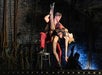 Dancers perform a dramatic lift on stage, with one sitting on a chair holding the other, who is in fishnet stockings, against a dark brick wall backdrop.