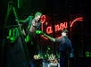 Two actors perform on stage; one stands on a ladder holding a bottle, handing it to the other. A neon sign reading “L’amour” glows red in the background amid green lighting.