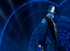 A man in a top hat and dark coat stands on stage under blue lighting, with an ornate, circular backdrop behind him.