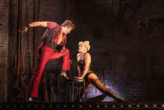 Two actors perform on stage; one stands with a foot on a chair, leaning toward the other, who sits on the chair in a dramatic pose, against a backdrop of ropes and a brick wall.