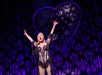 Performer in a black and white corset costume sings on stage in front of a large decorative heart and purple patterned backdrop.