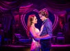 A couple stands closely on stage, gazing into each other's eyes. The background features a heart-shaped decoration with a purple and pink color scheme.