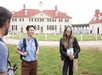 Two people stand and talk outside a large historic building with red roof and white walls, while a third person faces them partially out of frame.