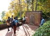 A group of people listen as a guide gestures toward a small wooden door set in a brick wall outdoors, surrounded by trees in autumn.