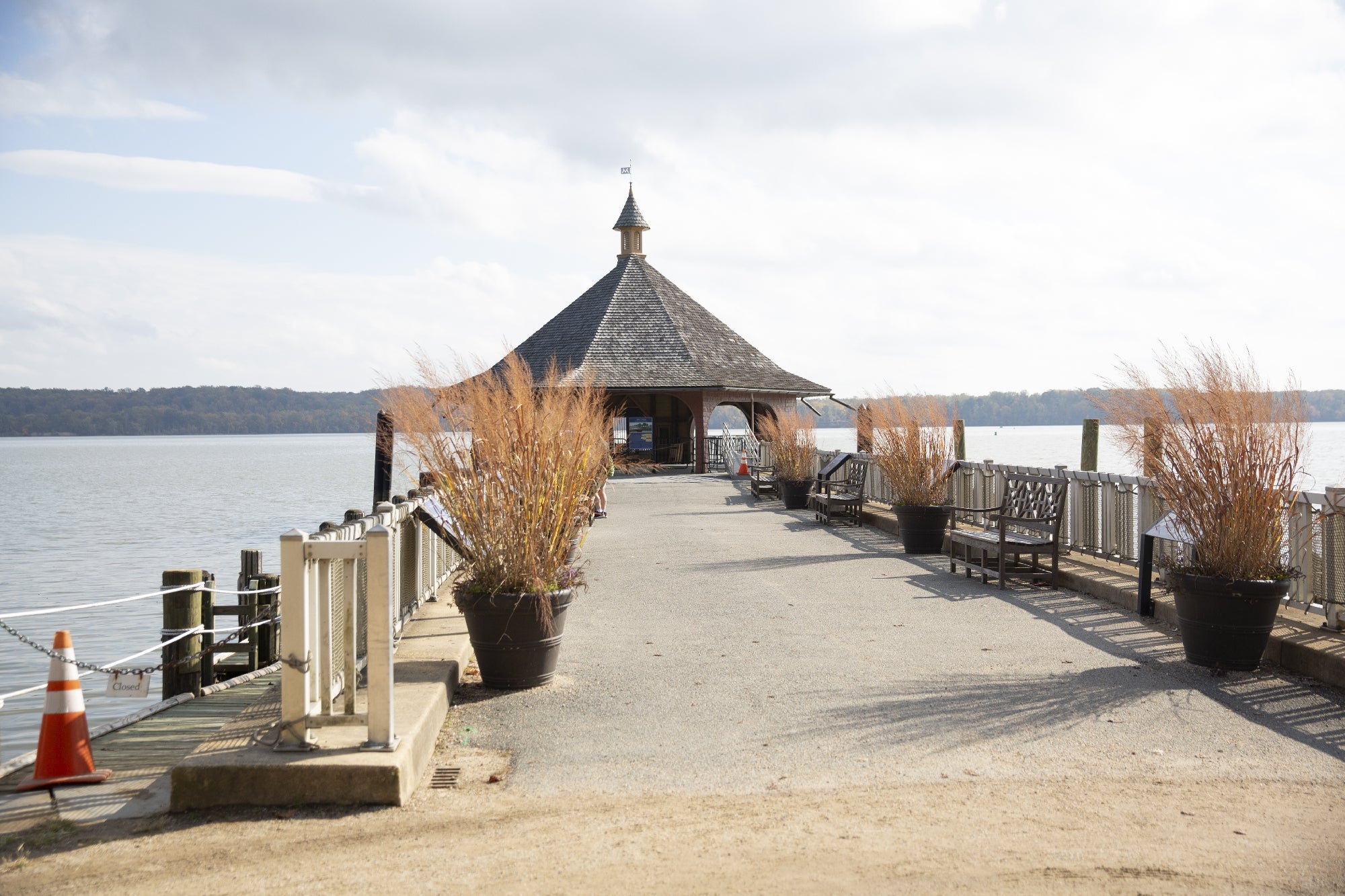 A wooden gazebo sits at the end of a pier over a calm lake, flanked by benches and large potted grasses under a partly cloudy sky.