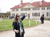A woman wearing sunglasses gestures and speaks in front of a historic building with a red roof and white walls, likely guiding a tour.