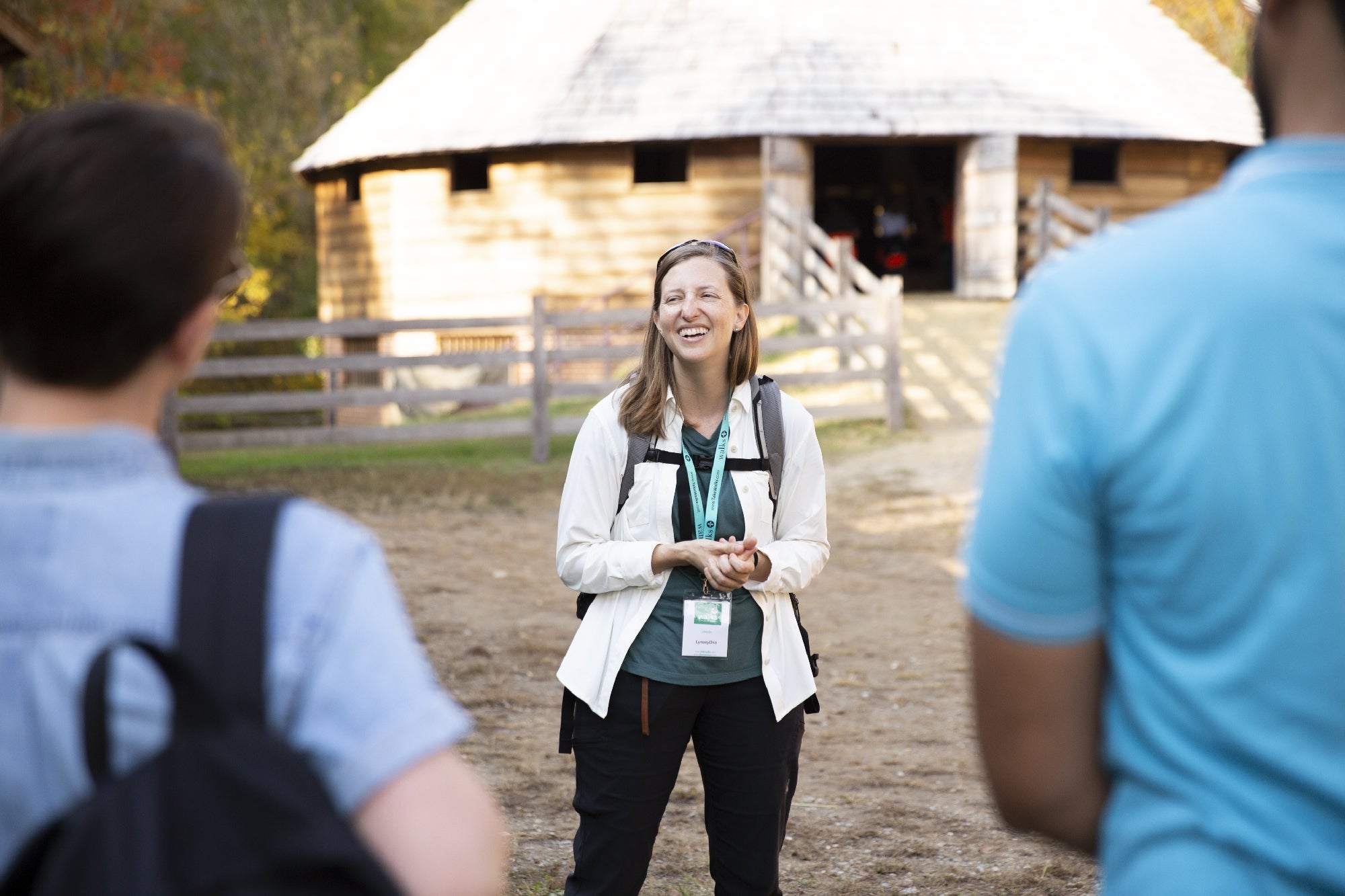 A woman with a name badge speaks to two people outdoors in front of a rustic wooden building and split-rail fence.
