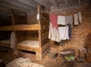 Wooden bunk beds and hanging laundry inside a rustic brick-walled room with baskets of vegetables and simple furnishings on a brick floor.