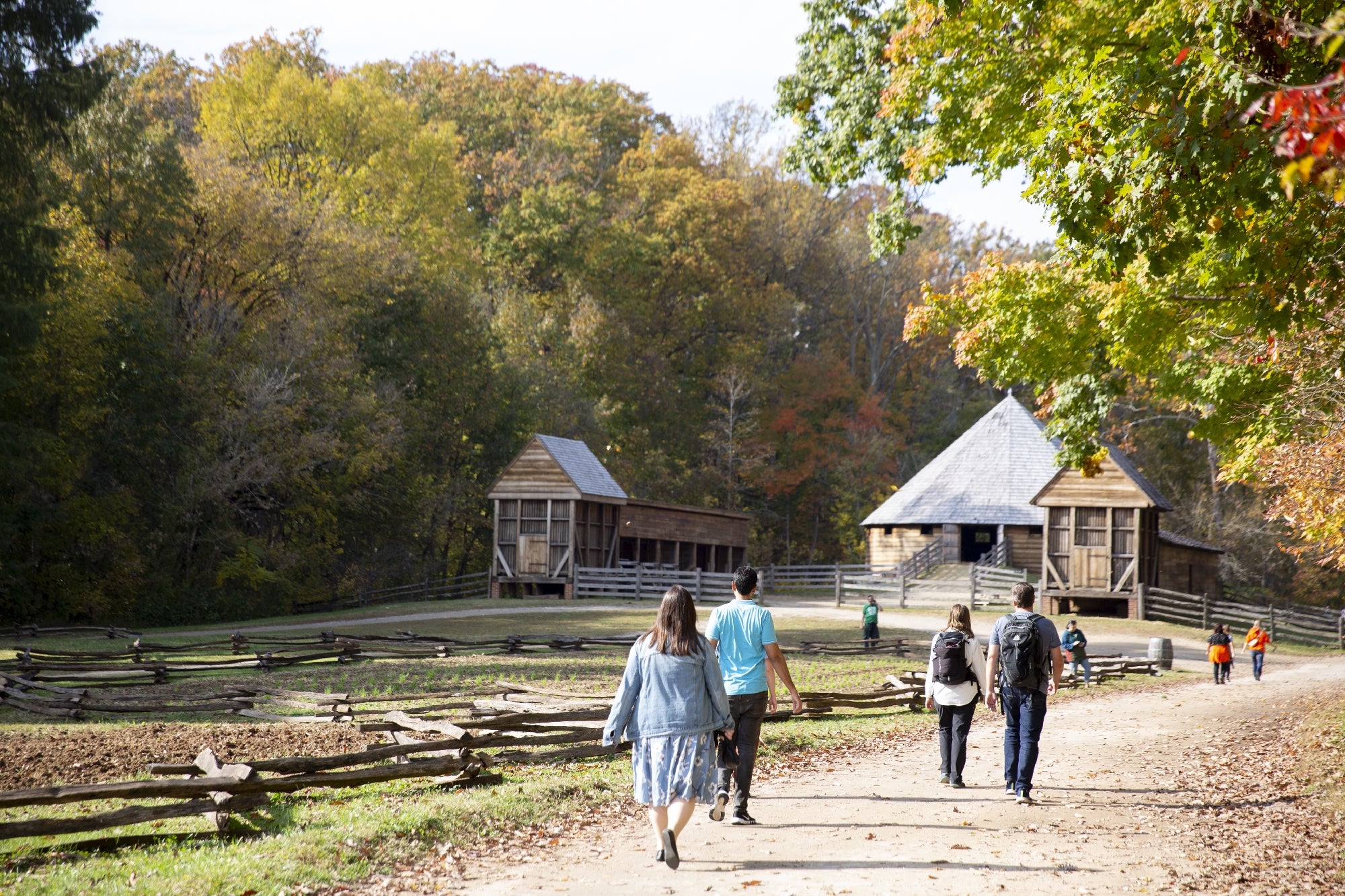 People walk along a dirt path toward historic wooden buildings surrounded by a split-rail fence and autumn trees.