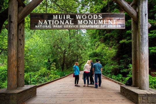 A group of people walking of a wooden bridge leading into the woods with the wooden arch sign for the Muir Woods National Monument behind them.