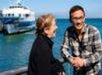 Two people stand on a dock near a boat, smiling and laughing with the ocean in the background.