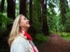 Person in a white jacket and red scarf stands smiling, looking up among tall trees in a forest.