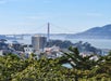 View of the Golden Gate Bridge in San Francisco with buildings, trees, and hills in the foreground under a clear sky.