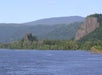 A wide river flows past green, forested hills and rocky cliffs under a clear sky.
