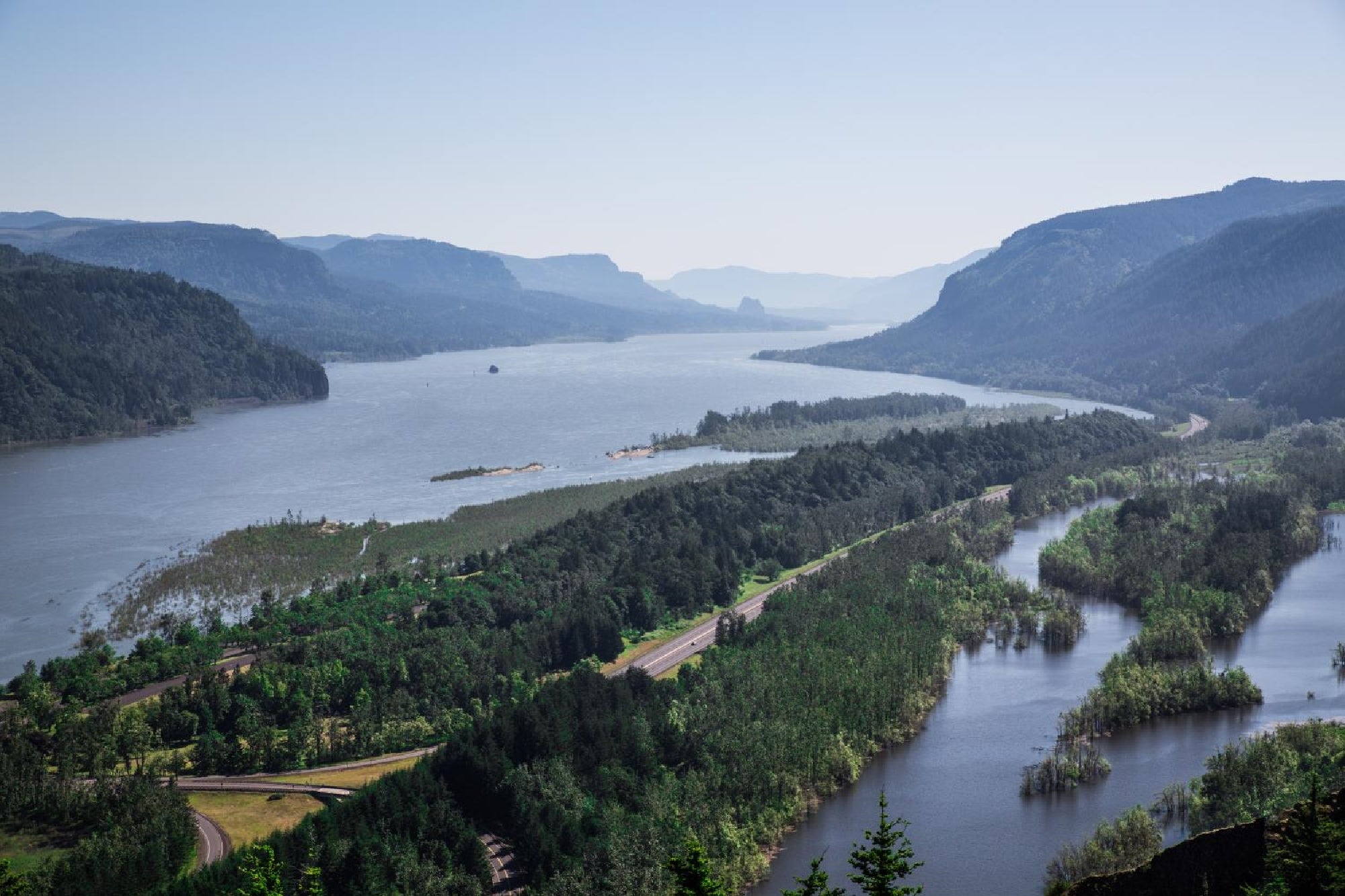 A wide river flows between forested mountains under a clear sky, with multiple green islands and a road running alongside the water.