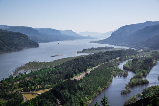 A wide river flows between forested mountains under a clear sky, with multiple green islands and a road running alongside the water.