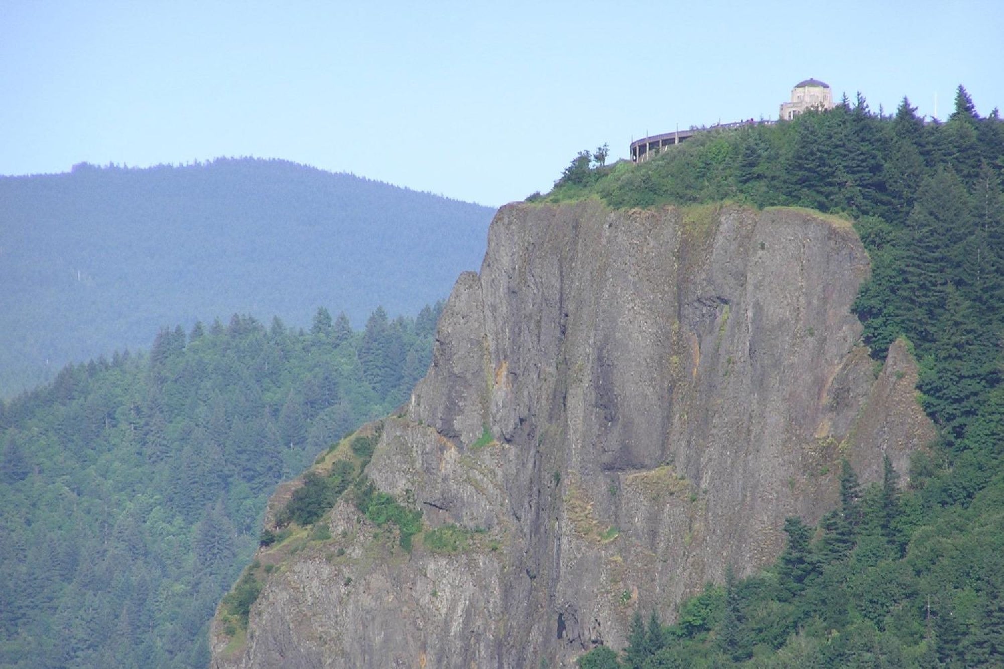 A rocky cliff covered partly with trees, with a building located at the top, set against a backdrop of forested hills and a clear sky.