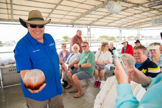 A man in a blue shirt and hat holds a large round object while smiling, as a seated group of people watch and someone takes a photo on a boat.