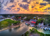 Aerial view of a riverside town at sunset, with boats docked along the shore, marshy areas, buildings, and a vibrant sky with scattered clouds.
