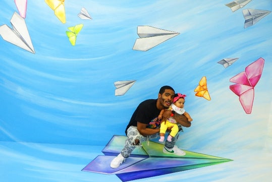 A man and a baby pose on a large illustrated paper airplane, surrounded by colorful paper airplane drawings on a blue-themed background.