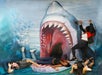 Four people pose in front of a large, realistic shark mural; two appear inside its open mouth, while the others lie on the ground as if being attacked.