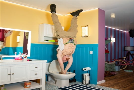 A man performs a handstand on top of a toilet in a brightly colored bathroom, with his hands inside the toilet bowl and feet touching the ceiling.