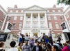 A crowd of people gathers on the steps outside a large, multi-story brick building with white columns and signs on either side of the entrance.