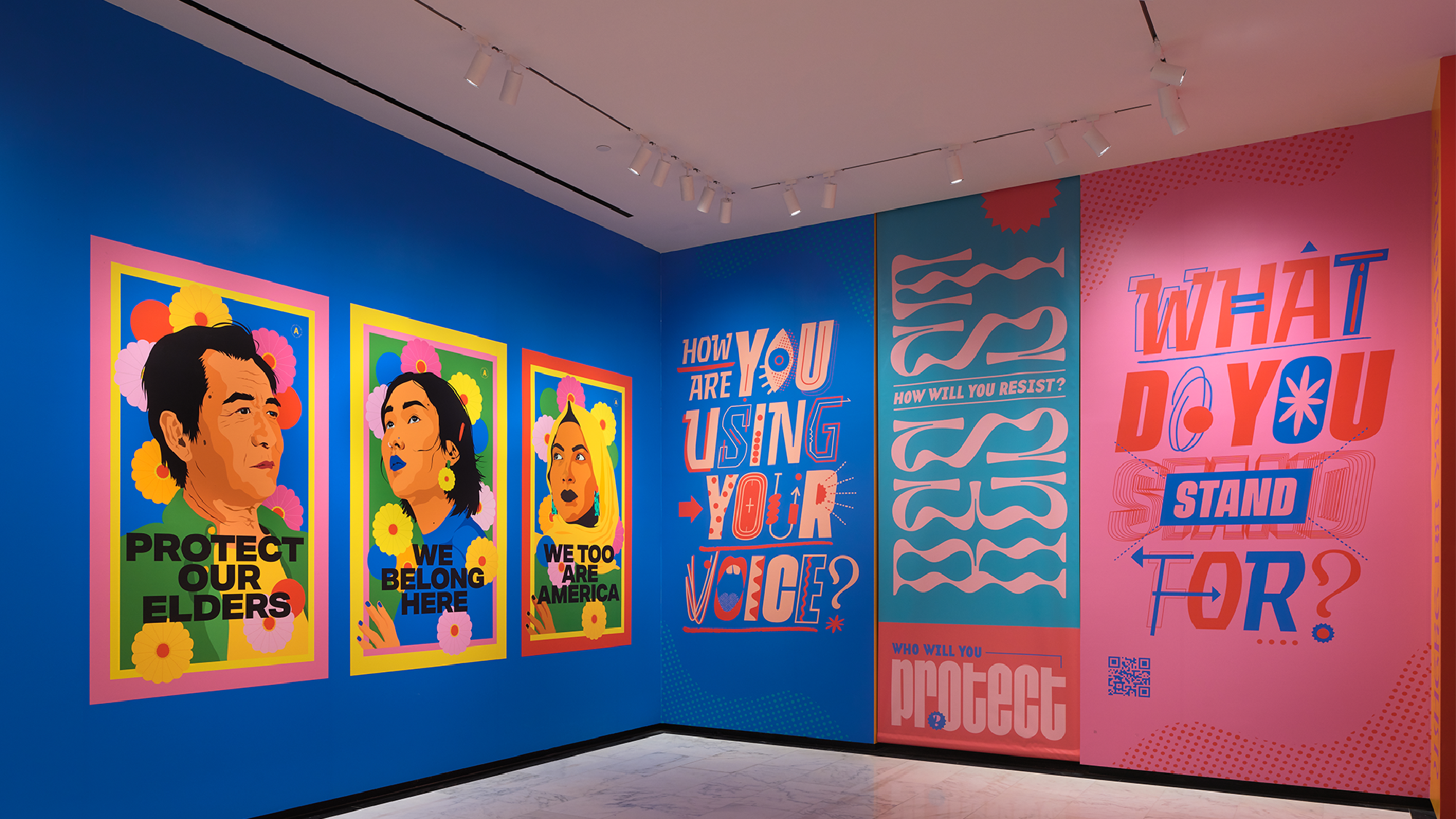 Colorful art exhibit features posters with illustrated portraits and bold text messages, including “PROTECT OUR ELDERS,” “WE BELONG HERE,” and “WHAT DO YOU STAND FOR?” on blue and pink walls.
