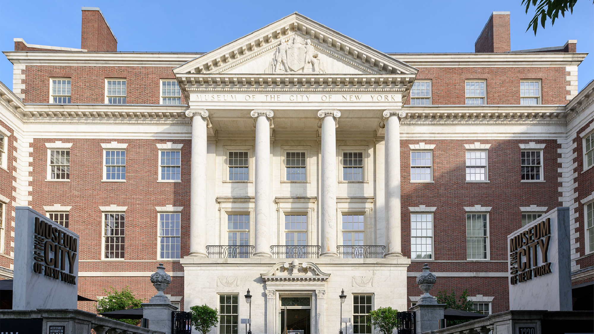 Front view of the Museum of the City of New York, featuring a red brick facade, tall white columns, and two entrance signs.