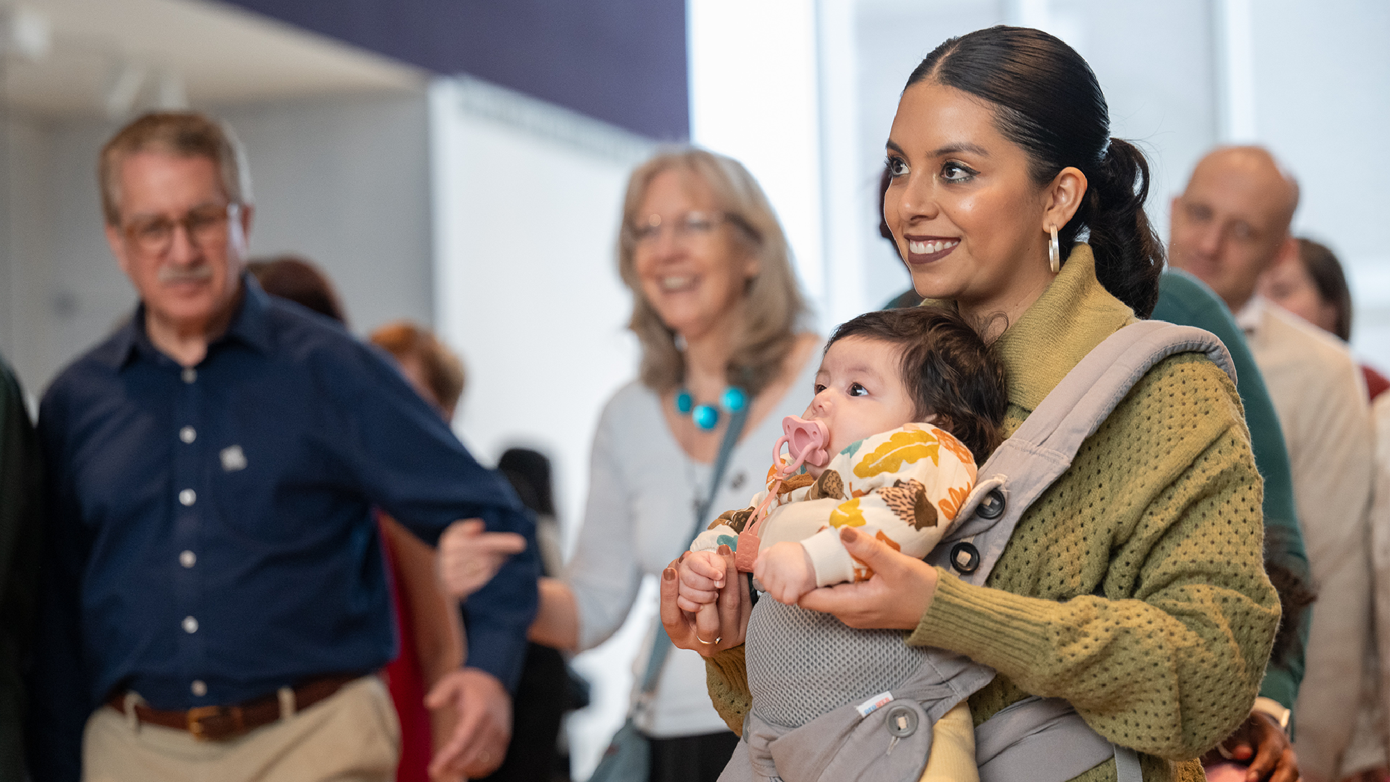 A woman in a green sweater holds a baby in a carrier, surrounded by smiling adults in a brightly lit indoor setting.