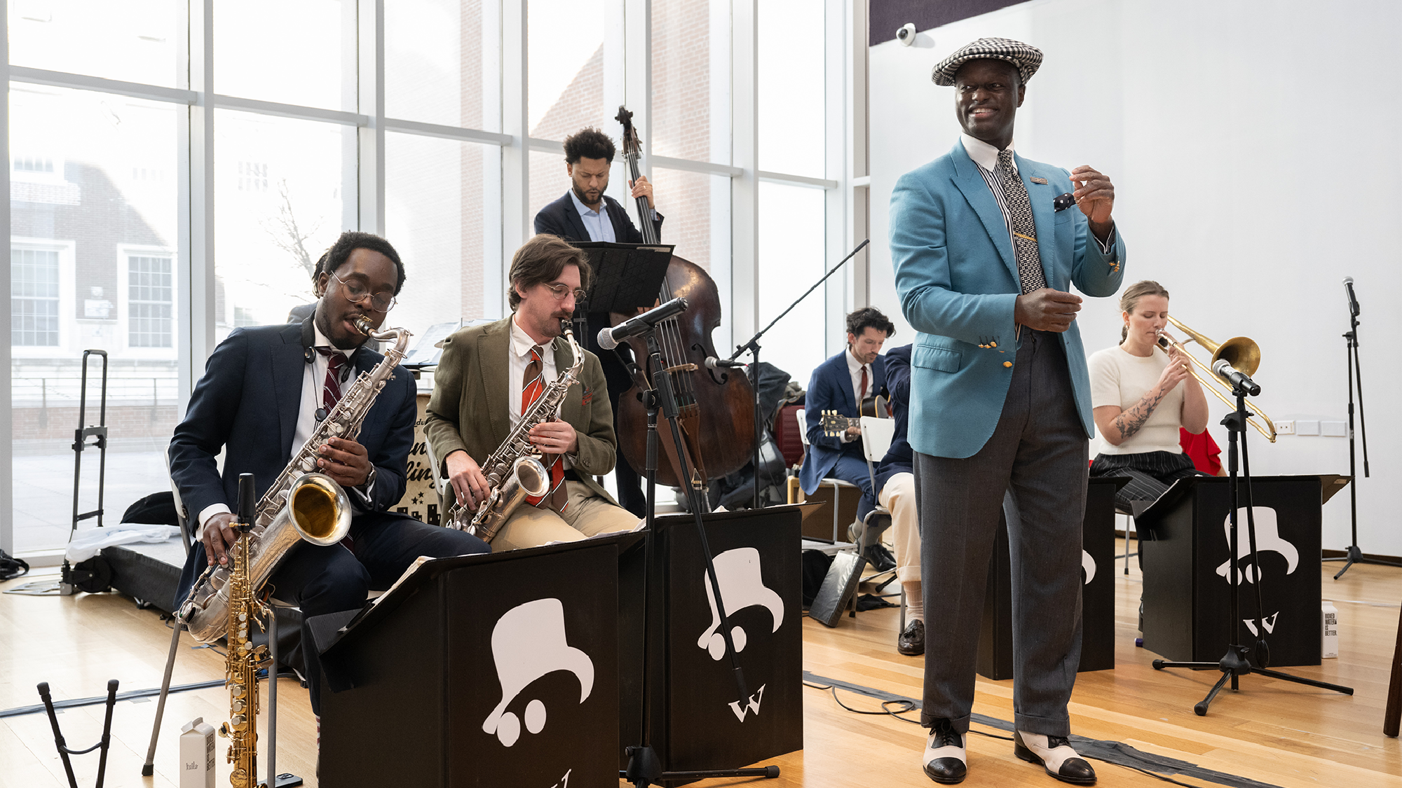 A jazz band performs indoors with saxophones, a double bass, trombone, and a vocalist in a blue blazer standing at the front.