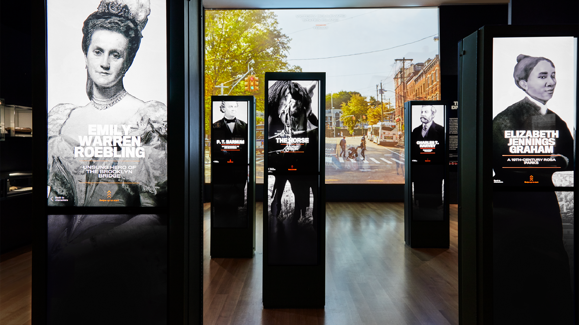 Exhibit displays with images and text about historical figures, including Emily Warren Roebling and Elizabeth Jennings Graham, are shown in a modern museum space with a city street photo in the background.
