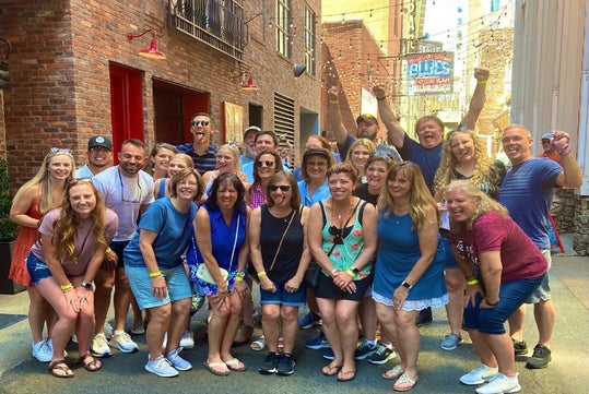 A group of people poses smiling in an alley. They are casually dressed, and the setting includes brick walls and signage in the background.