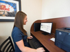 A woman in a blue shirt uses a desktop computer at a wooden desk, next to a printer. A framed picture with "Wolves" is partially visible on the wall.